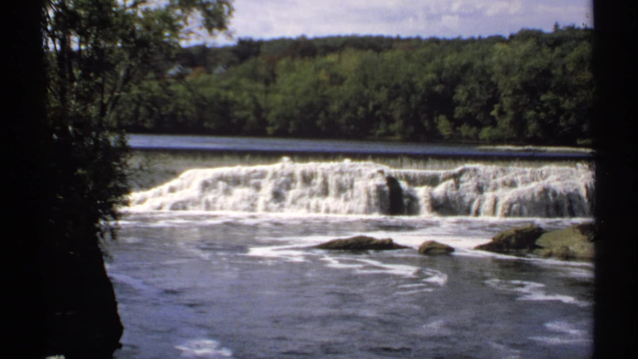 River water flowing over a low dam with trees and rocks around the banks.