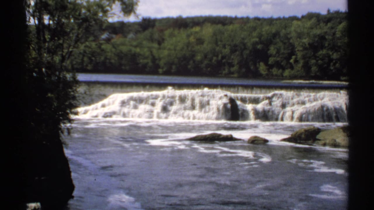 Wide river cascade seen between dark tree trunks with woods beyond.