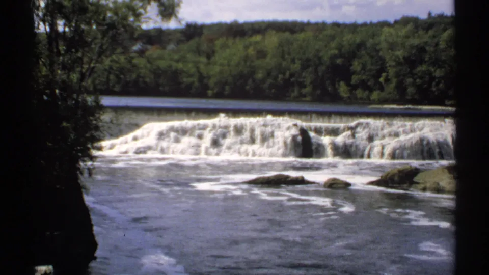 1971 Maine 8mm view of a river cascade framed by dark tree trunks, with a wooded shoreline across the water.