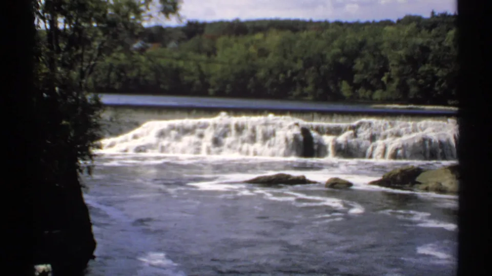 1971 Maine 8mm frame of water spilling over a low dam, with rippled river surface, rocks, and dense trees behind it.