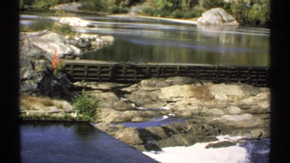 1971 Maine 8mm frame showing a rocky riverbank, calm pool, small wooden crossing, and foam where water moves over rocks.