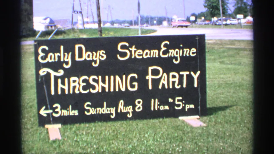 1971 Minnesota 8mm frame showing a roadside sign for the Early Days Steam Engine Threshing Party, with cars and open grass behind it.