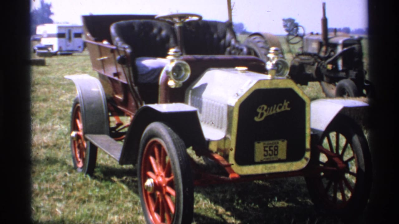 1971 Minnesota home-movie frame showing a vintage Buick on grass, with red wheels, brass-style lamps, a white camper, and equipment behind it.