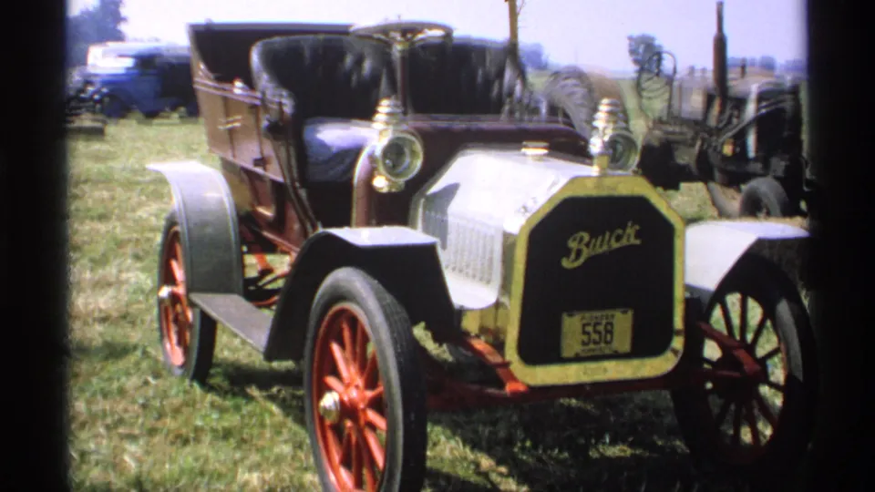 Home-movie view of the antique Buick display in Minnesota, with its white hood, red wheels, black interior, and old equipment at the edge of frame.
