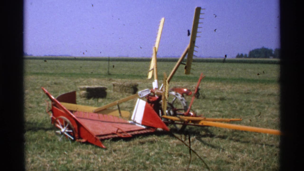 Red and yellow hay rake resting in a grassy field under a blue sky.