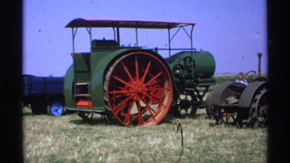 1971 Minnesota 8mm frame of a green steam tractor with red spoked wheels, canopy and a small person near the right side.