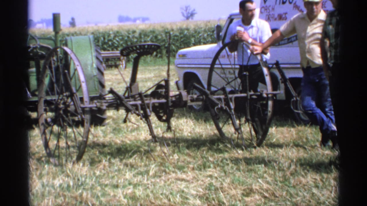 Antique farm implement in a grassy field with men and a white pickup behind it.