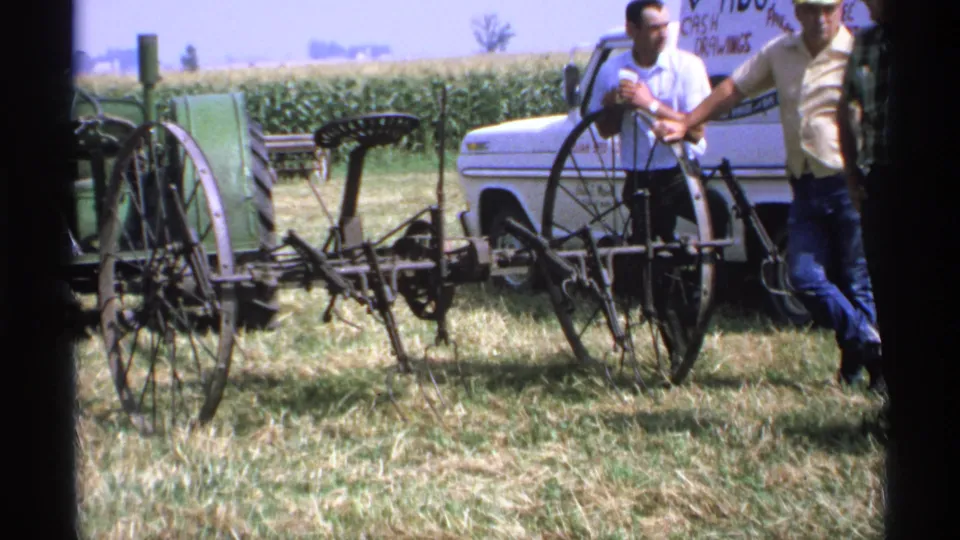 1971 Minnesota 8mm frame showing an antique wheeled farm implement displayed outdoors with men and a white pickup nearby.