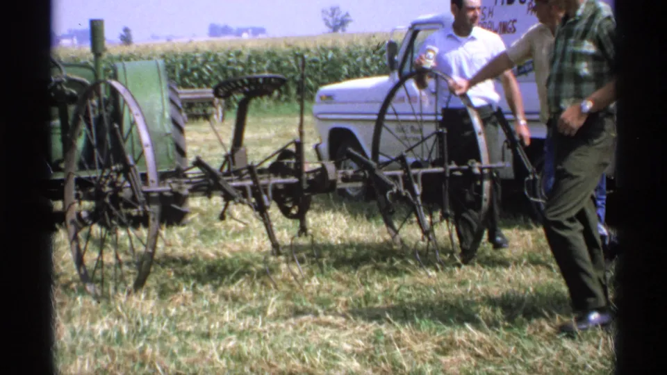A man in a plaid shirt and others stand by an antique farm implement at a 1971 Minnesota outdoor display.