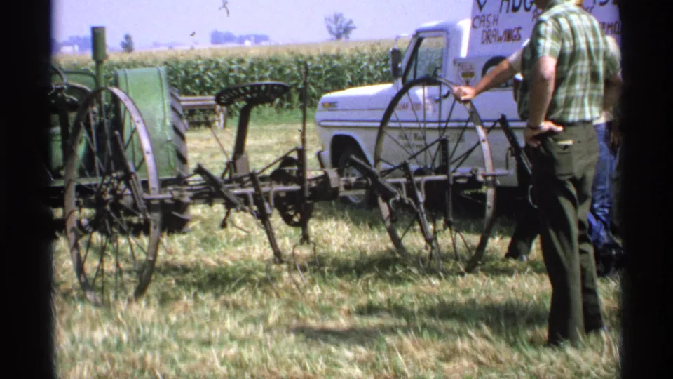 1971 Minnesota frame with a man in a green plaid shirt viewing an antique farm implement near a pickup and crop field.