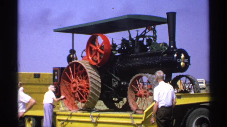 A large black steam tractor with red wheels is displayed on a yellow trailer in 1971 Minnesota home-movie footage.