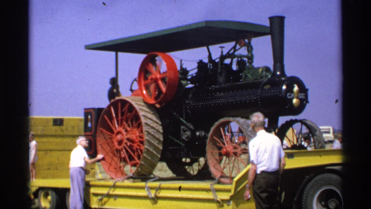 Black steam traction engine with red wheels on a yellow flatbed as two men stand nearby.