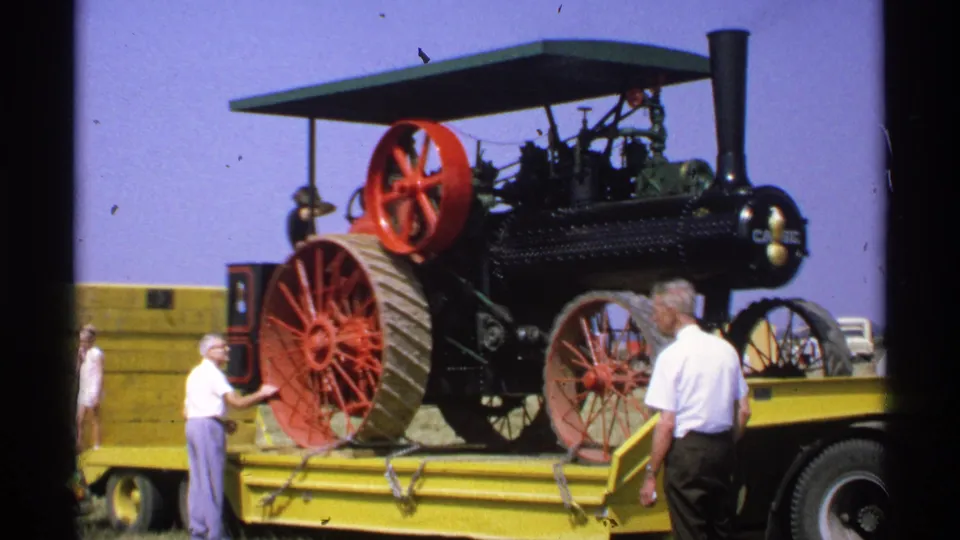 1971 Minnesota frame of an antique Case steam traction engine on a yellow flatbed, with men positioned around the machinery during unloading.