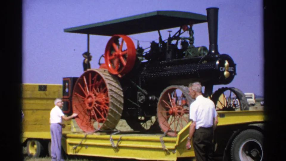 1971 Minnesota home-movie still showing men beside a Case steam traction engine secured on a yellow flatbed trailer in an open field.