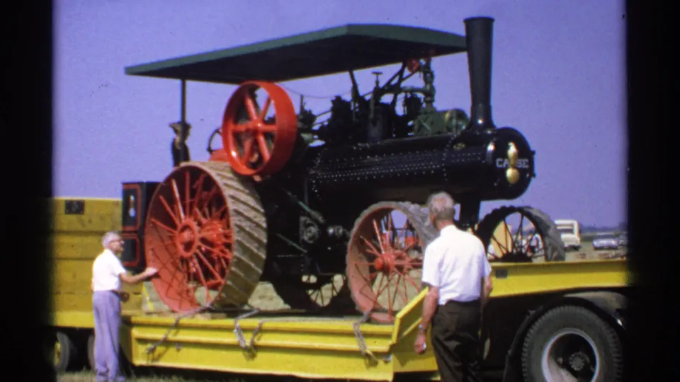 1971 Minnesota frame showing a restored-looking Case steam traction engine on a yellow flatbed with men nearby during transport or unloading.
