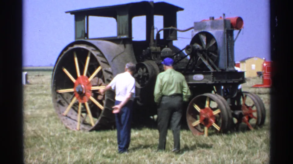1971 Minnesota home-movie frame showing two adult men examining the rear of a large vintage tractor or steam machine at a fairground.