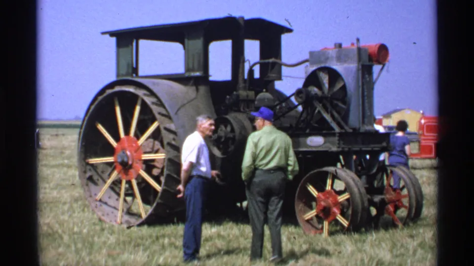 1971 Minnesota frame showing adults around a vintage tractor's rear and drive components during an outdoor machinery display.