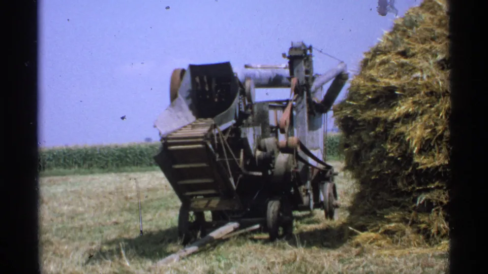 Minnesota 1971 8mm frame showing a large belt-driven farm machine beside a haystack, with a field and corn rows in the background.