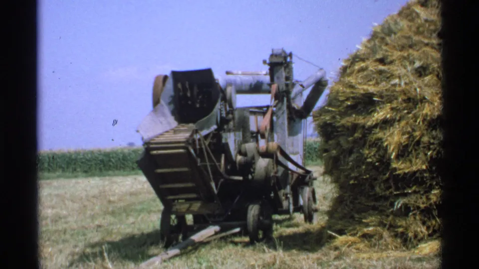 Minnesota 1971 8mm frame showing the vintage farm machine from a distance beside hay, with cornfield and sky in view.