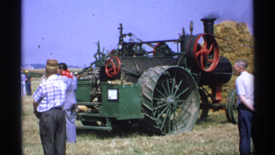 1971 Minnesota home-movie frame of adult men observing a green steam-powered agricultural machine with large wheels and red flywheels in a rural field.