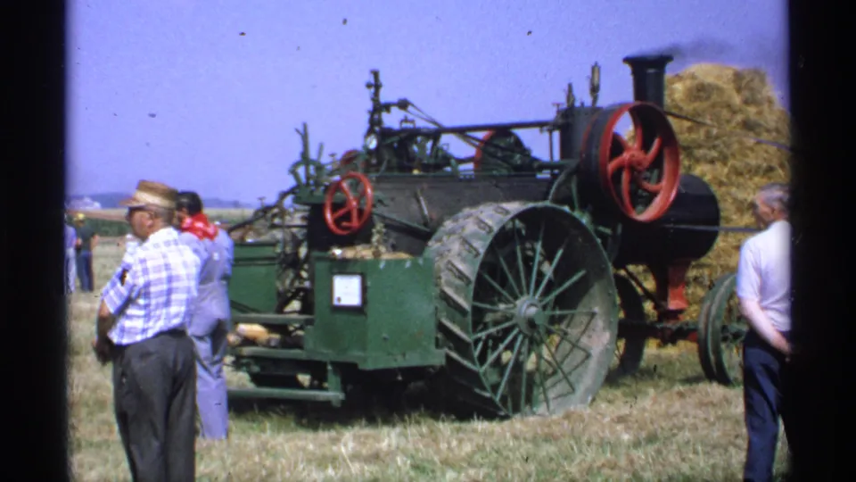 1971 Minnesota frame showing several adults watching a green vintage agricultural machine, with a straw-hatted man in plaid closest to the camera.