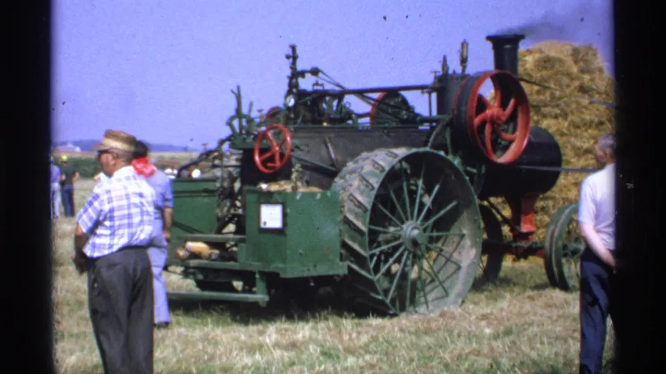1971 Minnesota 8mm frame of a green steam-era farm machine with large spoked wheels, red flywheels, and adult men standing beside it.