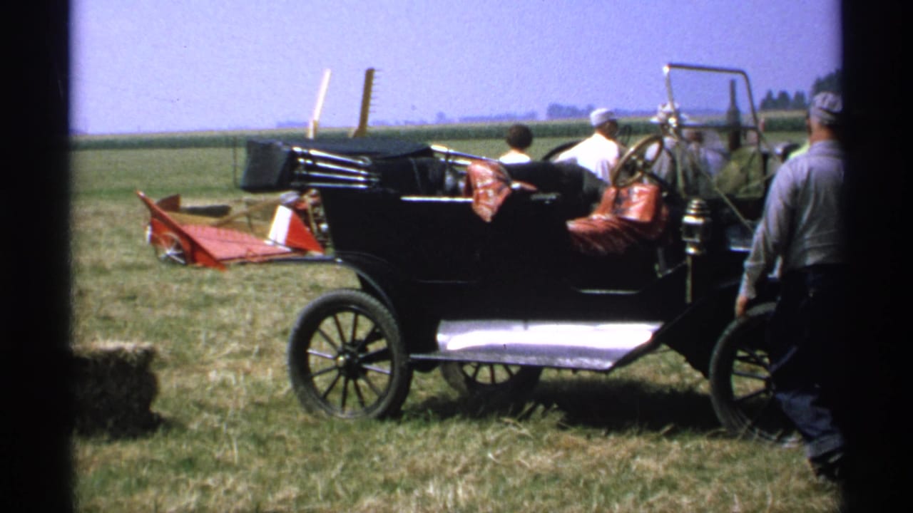 1971 Minnesota 8mm frame showing a black early automobile in a field with adults gathered around it and rural equipment in the background.