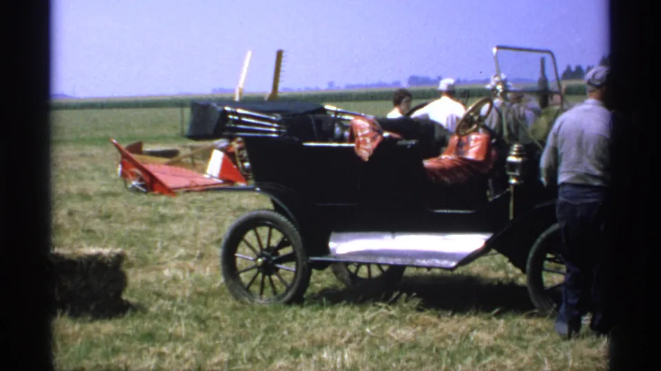 1971 Minnesota home-movie frame with a black vintage automobile, adults around the passenger area, and open farmland beyond.