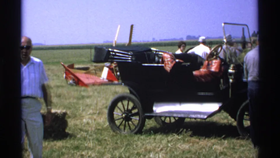 1971 Minnesota 8mm frame showing a foreground adult in sunglasses passing a black antique car in a rural field setting.