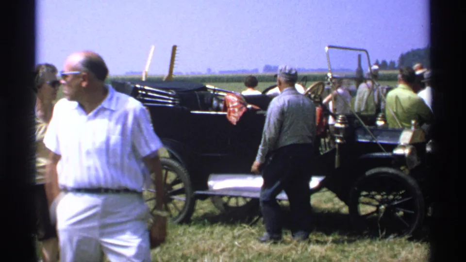 1971 Minnesota home-movie frame with adults moving around a black antique automobile at a rural outdoor gathering.