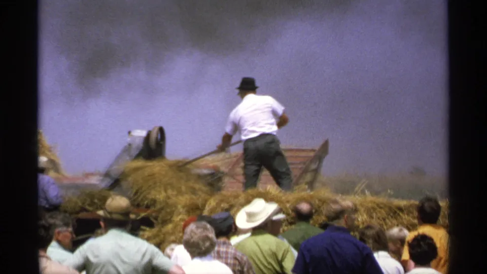 A man continues moving hay toward a conveyor while dust or smoke drifts behind him and spectators watch nearby.