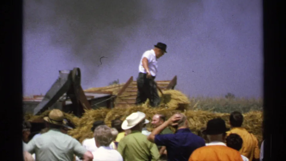 A worker pauses or shifts position on a hay pile beside conveyor machinery while the crowd remains in the foreground.