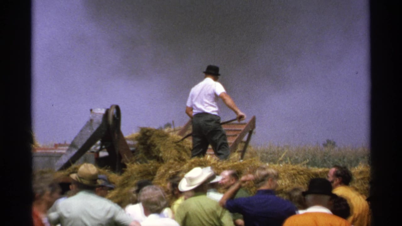 1971 Minnesota 8mm frame of a man in a white shirt and dark hat working near hay as spectators watch a farm demonstration.