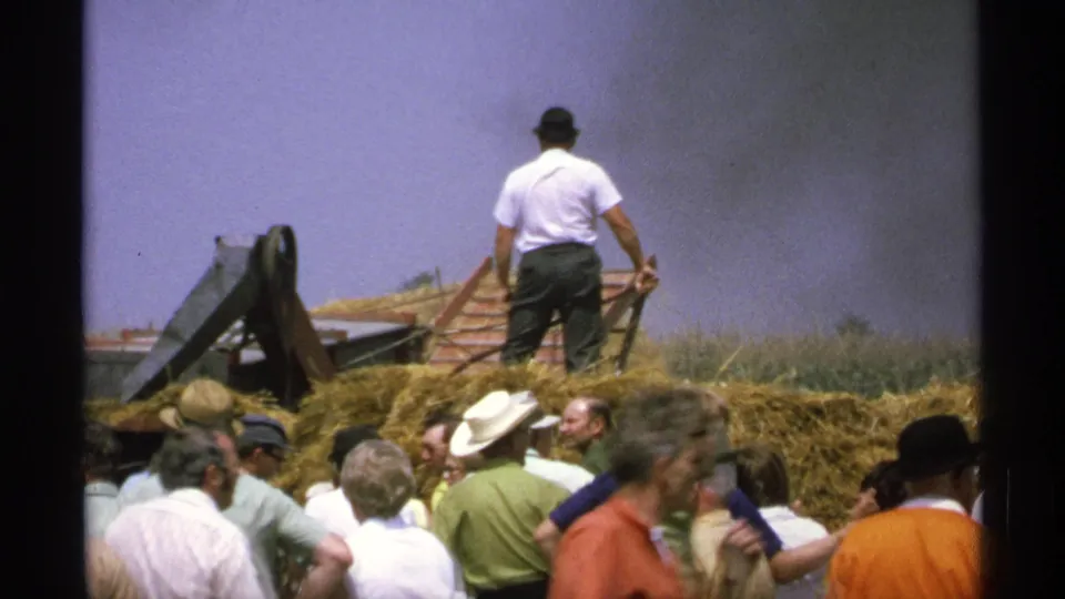 The operator faces away while handling equipment on a hay pile, with a crowd in summer clothing watching in Minnesota archival footage.