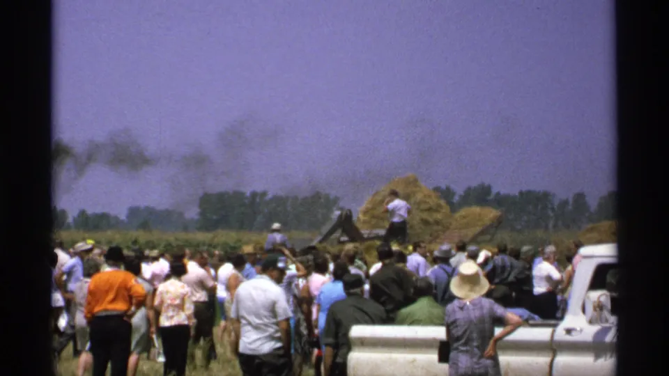 Wide 8mm view of spectators gathered in a field as a man works near hay and farm machinery during a Minnesota demonstration.