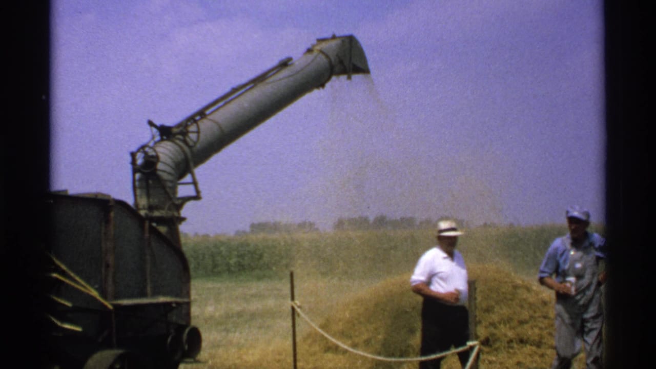 Hay-processing machine ejecting hay into a pile while two men stand nearby in a field.