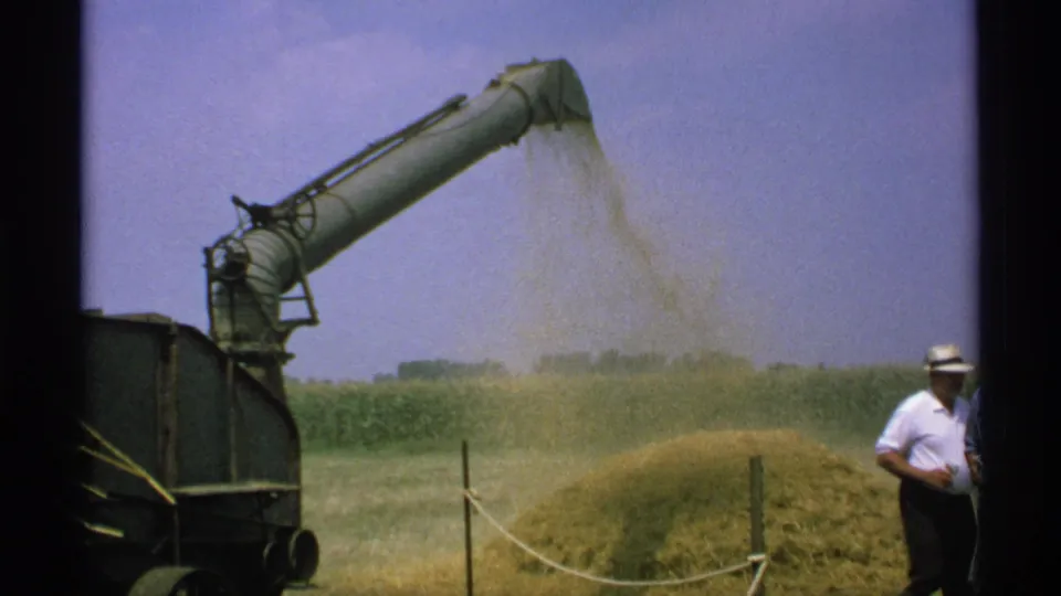 The hay chute continues ejecting material into a growing pile while a man in a white shirt and hat stands near the machinery.