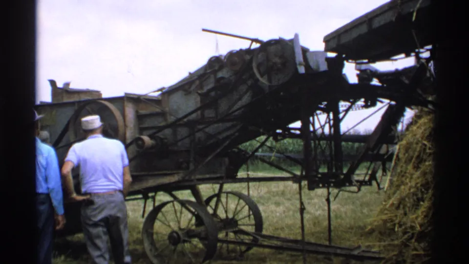 A side view shows the wheeled hay-processing machine, belts, frame supports, hay pile, and several men gathered in the field.