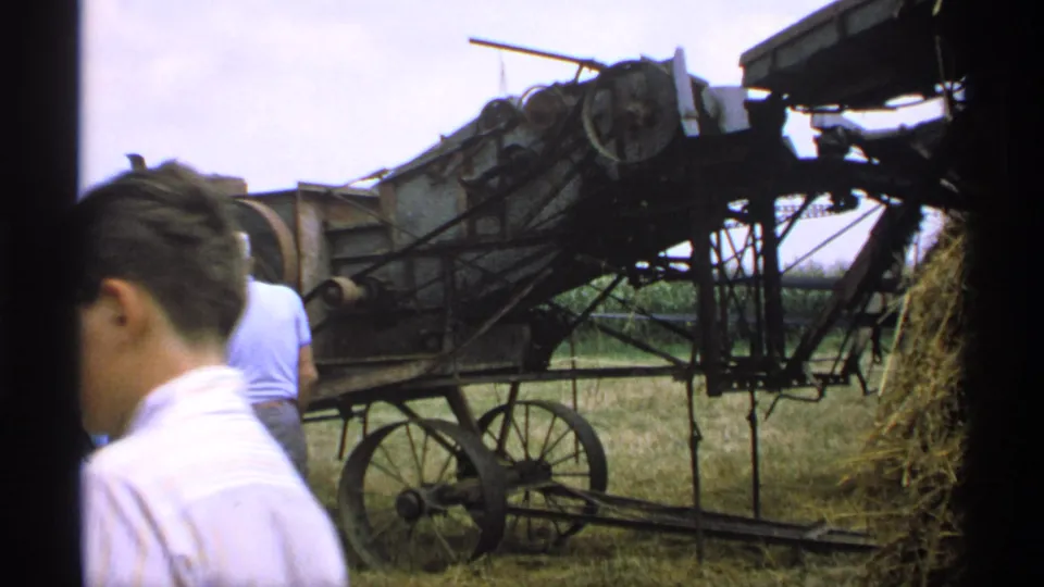 A young male figure crosses the foreground while the wheeled hay-processing machine and hay pile remain visible in the Minnesota field.