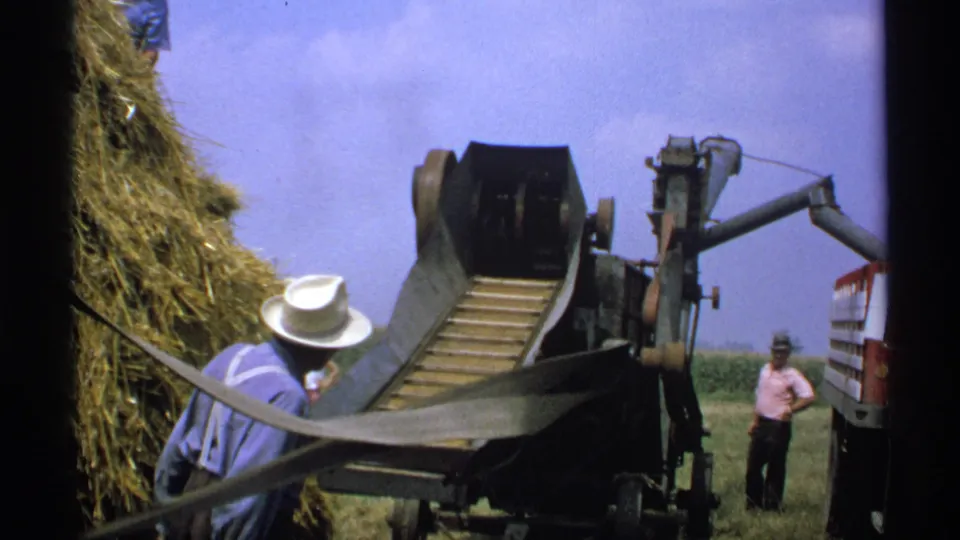 A Minnesota farm worker in a white hat stands near the baler chute while loose hay is fed toward the machine.