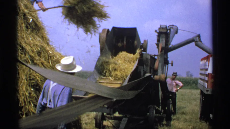In a rural Minnesota field, hay moves up the conveyor while one farmer works close to the baler and another man stands near the truck.