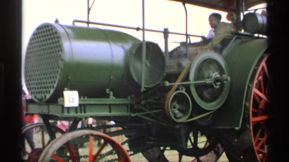 Close 1971 Minnesota 8mm view of the green steam tractor boiler, exposed belt drive, flywheel, and operator area.