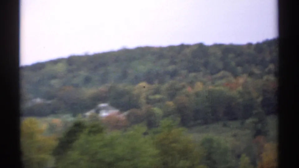 1971 New Hampshire home-movie frame showing a wooded hillside, pale sky, dark borders, and a small light-colored structure in the valley.