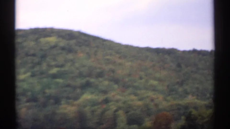 1971 8mm New Hampshire landscape frame with a green mountain slope, dense treetops, pale sky, and dark window-like edges.