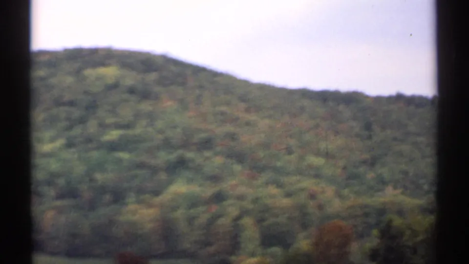 1971 New Hampshire 8mm frame showing a wooded mountain ridge with green canopy, faint autumn color, pale sky, and dark edge framing.