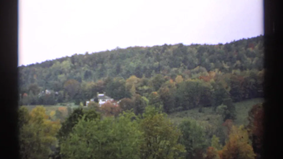 1971 New Hampshire home-movie frame showing a valley field, distant white houses, and a forested hillside in soft 8mm color.