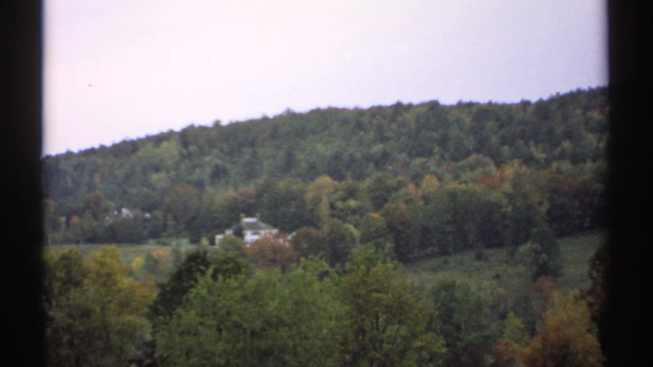 1971 New Hampshire countryside view with a white house set among trees below a green wooded hill.