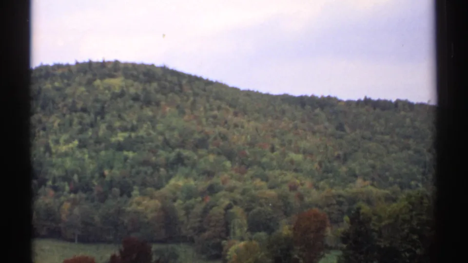 1971 8mm view of a wooded New Hampshire hillside with early autumn color, framed by dark vertical window edges.
