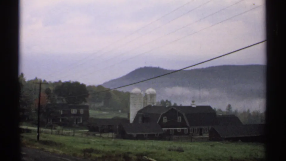1971 New Hampshire rural farm view with a red barn, white silos, pasture, overhead wires, and mist over low hills.
