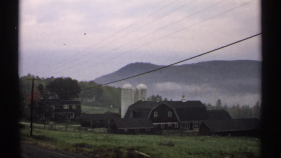 New Hampshire farm buildings and two white silos appear below low fog and wooded hills in this 1971 home-movie frame.